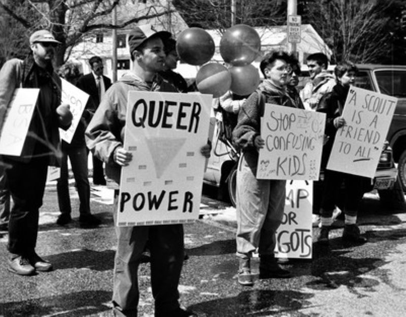 A photo from a protest against homophobia in the Boy Scouts of America. One protester is holding a sign that reads "Queer Power". Another holds a sign that says, "A Scout is a Friend to ALL."