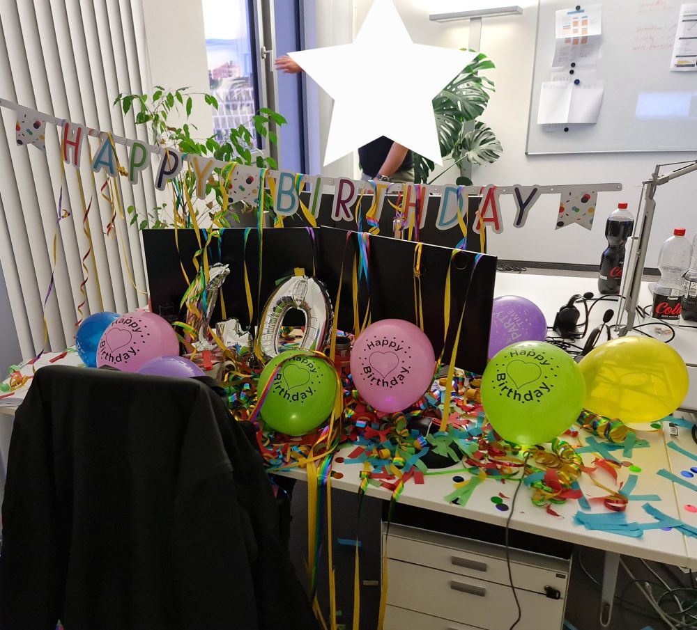 schreibtisch mit zwei bildschirmen 
Alles geht in buntem Lametta, luftballons und Konfetti unter.
Im Hintergrund ist ein  "Happy Birthday" Schriftzug