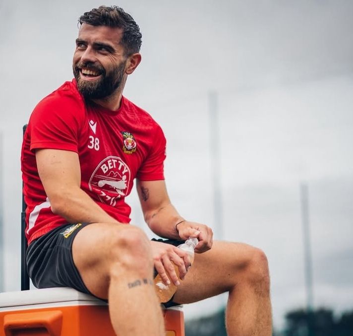 Wrexham AFC player Elliot Lee takes a break from training and sits on a cooler box while holding a bottle of liquid. He's looking back over his shoulder at something and has his trademark smile on his face. The skies are grey but Lee is bright in his red short-sleeve shirt and black shorts. 