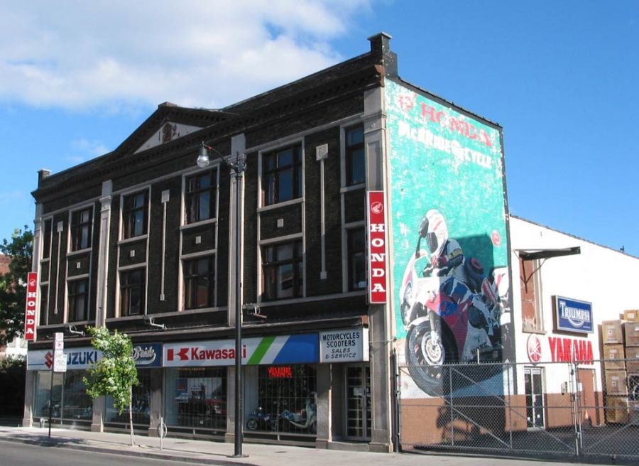 McBride Cycle, a 3-story brick building with a 2-story garage out back. Dealer has branding for Honda, Suzuki, Kawasaki, Yamaha, and Triumph, along with a 3-story mural of a rider on a Honda sport bike leaning into a corner.