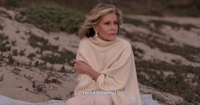 Jane Fonda sentada na praia com uma pelerine creme, com frio, e a legenda [wind blowing]