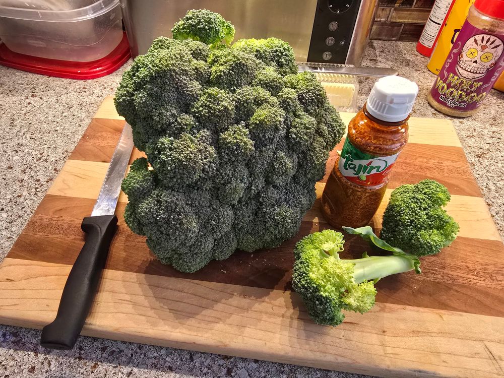 Broccoli on a cutting board with a bottle of tajin and a bread knife for size reference. 