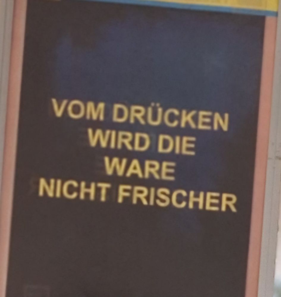Auf einem Schild steht mit gelben Großbuchstaben auf anthrazit-farbenem Untergrund zentriert geschrieben: VOM DRÜCKEN WIRD DIE WARE NICHT FRISCHER