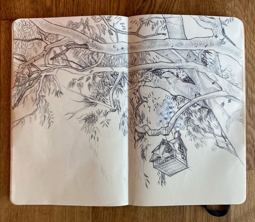 A sketchbook open on a wooden surface displays a closeup of tree, a small house hanging from one of its branches. A closer look of the house reveals a tiny car in the garage and smoke coming of of the chimney. 