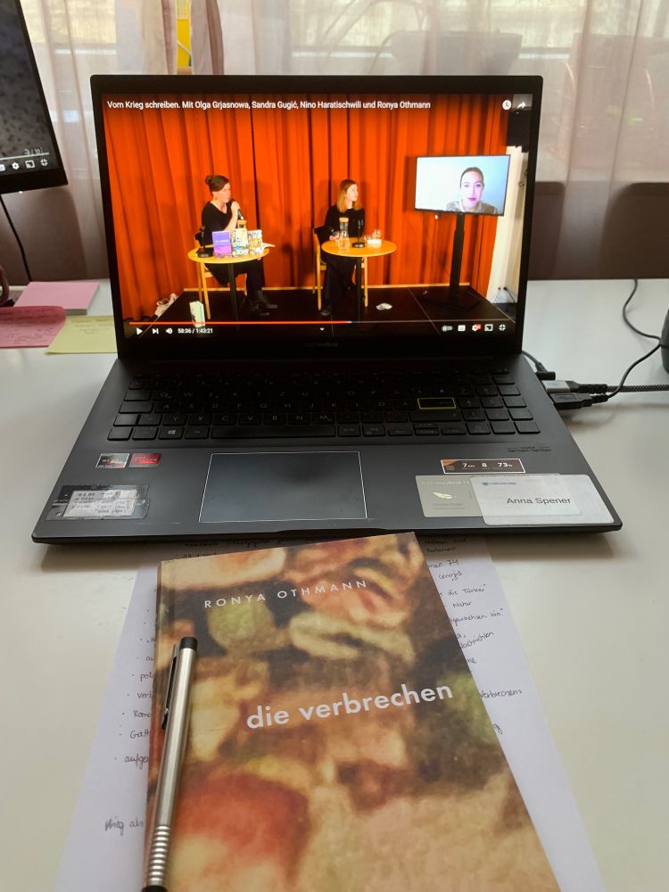 Foto eines Schreibtisches. Im Vordergrund liegt Ronya Othmanns „die verbrechen“, darunter ein Zettel mit Notizen, darauf ein Kugelschreiber. Im Hintergrund ist ein Laptop zu sehen mit einem geöffneten Youtube-Video „Vom Krieg schreiben. Mit Olga Grjasnowa, Sandra Gugić, Nino Haratischwili und Ronya Othmann“.