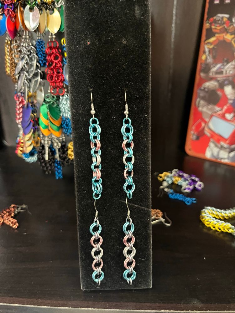 Two pairs of chainmail earrings, in the sky blue/pink/silver/pink/sky blue of the trans pride flag. Both in 2-2 chain; one with grey connectors, one with blue connectors.