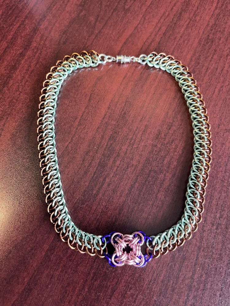A chainmail choker, mint and seafoam GSG on either side of a B^2 +1 “flower” in pink and purple. It lays on a wood grain table, with a silver barrel clasp holding it together.