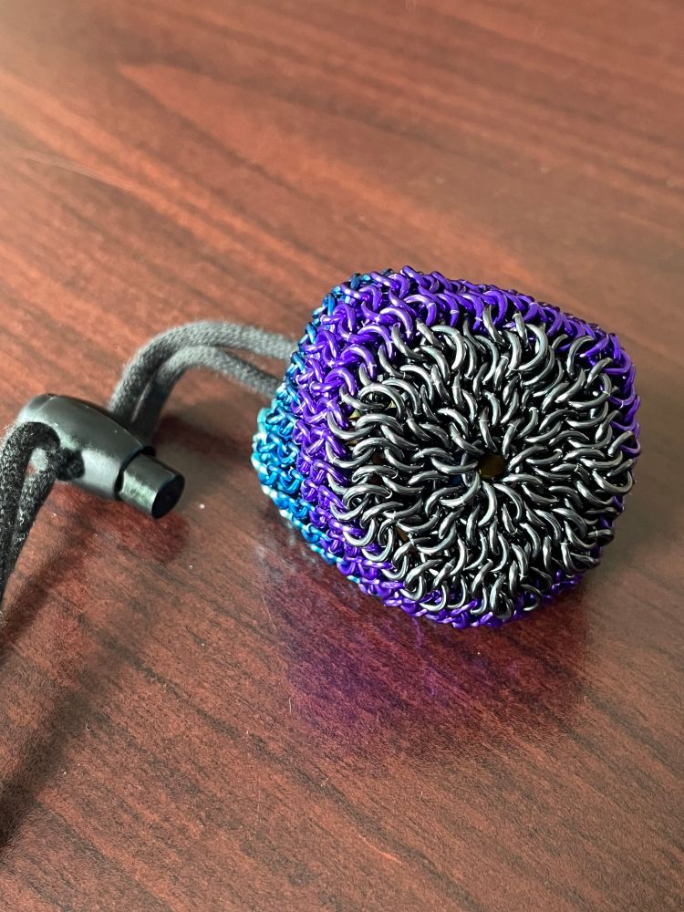 A small chainmail dice bag, filled, sitting on a wood-toned table. The bag is tilted with the bottom toward the camera, showing a black/grey array against the purple stripe. The cord and cord lock are black; the cord charm is not visible in this picture.