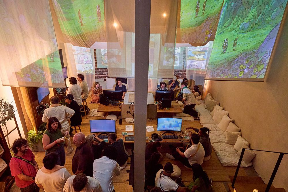 an overhead shot of a warmly lit teahouse room full of people -- some sitting on the floor, some standing and chatting. concept art is projected onto gauzey fabric hanging from the ceiling. there are 4 PC demo stations on short tables on the floor and 2 more standing PC demo stations on the side. the footage on the screen is censored.