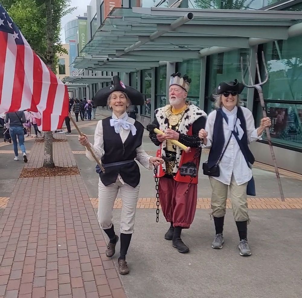 A trio at the Portland, OR No Kings protest. 2 are dressed in early US Colonial clothing, the center character is a king in chains. Person on the left holds a flag, person on the right a pitchpork.