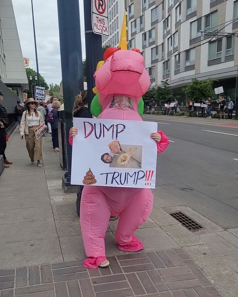 A pink unicorn at the No Kings protest in Portland, OR holding a sign that says "Dump Trump". Sign has an image of a pile of poop and a gold toilet.