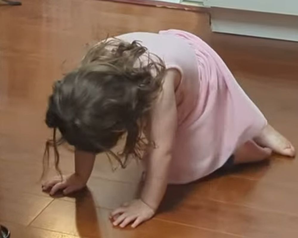 A toddler with messy hair and a pink dress crouched on a wooden floor.