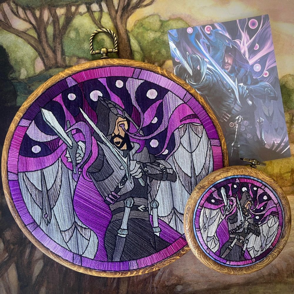 a photo showing the finished Lucanis embroidery next to a postcard print of the tarot card the pattern is based on, and also next to the smaller 3.5 inch embroidery mockup the artist completed previously