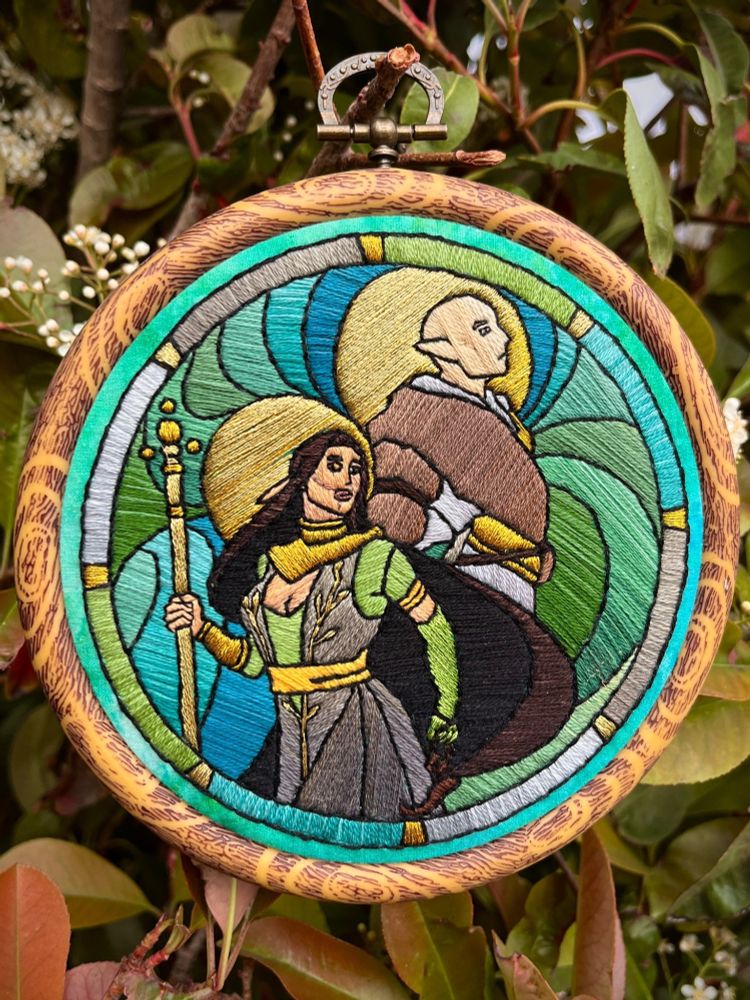 A 4.5 inch faux-wood embroidery hoop displayed hanging on a shrub. The embroidery is all done in satin stitch to mimic a stained glass or lineart art style,  featuring Solas and Lavellan from Dragon Age standing back to back with colorful swirls in the background. 