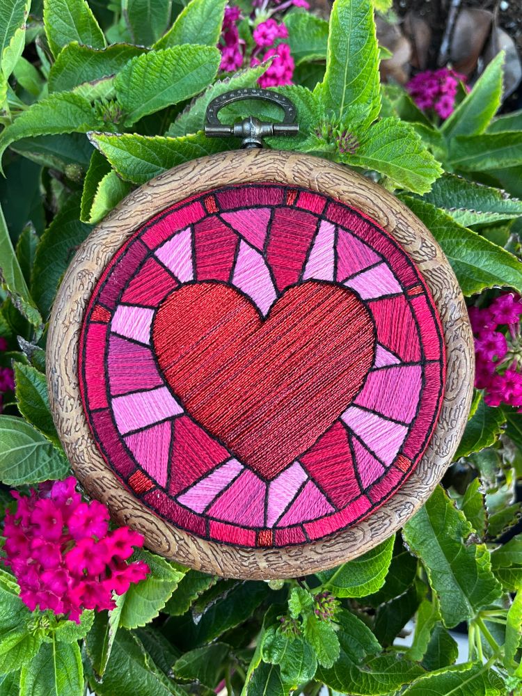 A photograph of a 3.5'' embroidery hoop resting on top of a pot with greenery and a few pink flowers. The hoop contains an embroidery of the "romance" icon art from Dragon Age: Inquisition of a heart surrounded by geometric shapes, mimicking the appearance of stained glass. The embroidery is done in all satin stitch to mimic the smoothness and shading of glass.