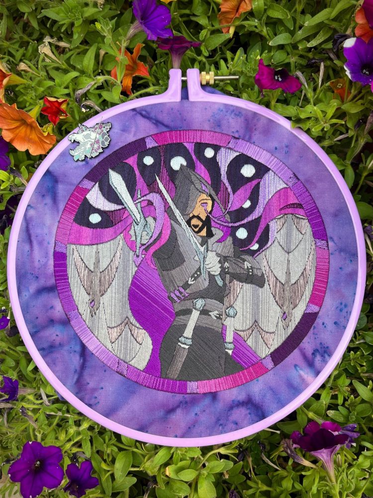 A photo of an embroidery hoop with a stitched design of the Lucanis tarot card from Dragon Age Veilguard in it. The pattern is entirely stitched, following a style mimicing stained glass. The hoop sits on top of a flower pot with green leaves and small colorful flowers in it.