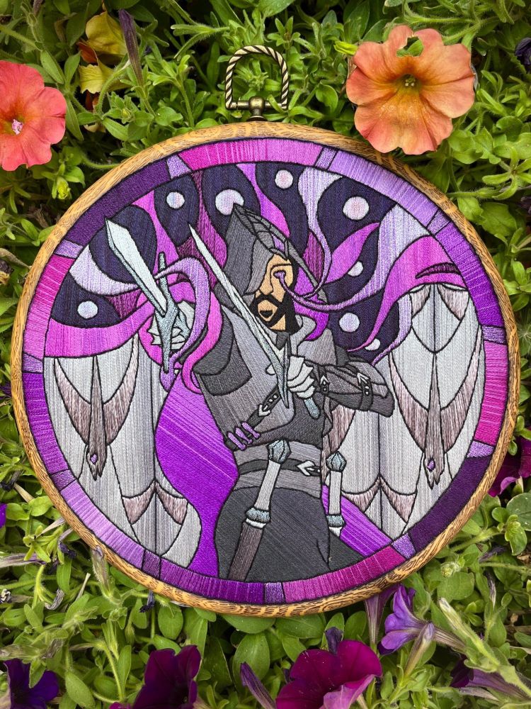 A top-down photo of an embroidery hoop resting on top of a pot of flowers. The stitching is of the character Lucanis from Dragon Age, and is done in a style to mimic the appearance of stained glass. The character stands in the middle holding two knives while the background is filled with purple swirls and grey geometric shapes resembling birds.