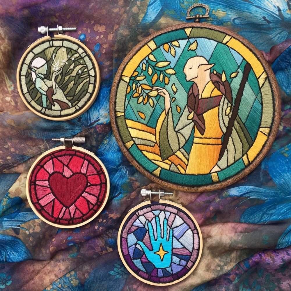a piece of fabric with 4 embroidery hoops on top of it, based off art from Dragon Age: Inquisition. There is a hoop with a red heart, a blue hand with a star in it, a green elf with magic swirls, and the character Solas as posed for his 'romance' tarot card the Hierophant. 