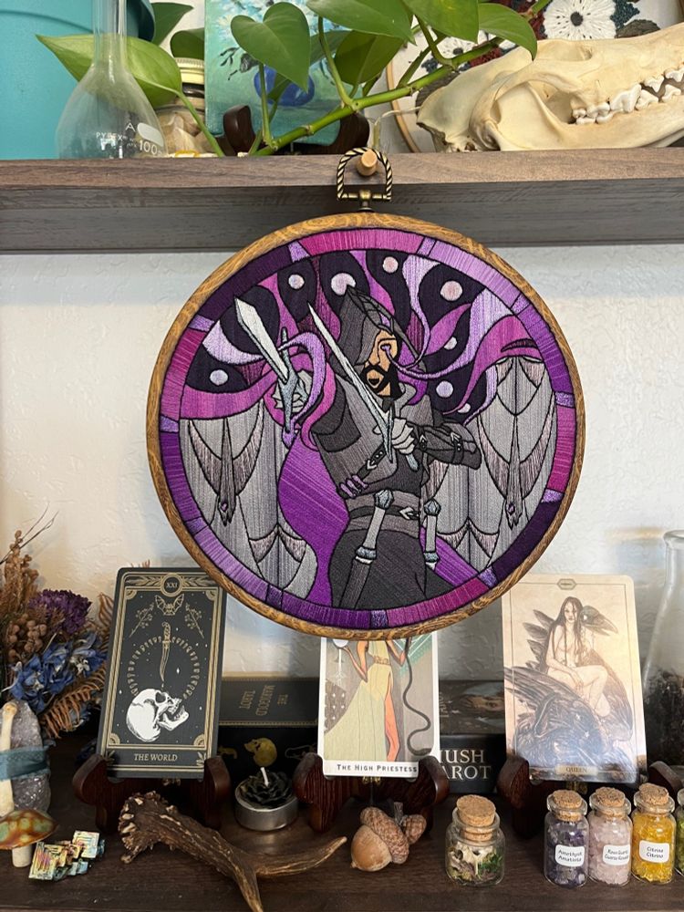 A photo of the finished Lucanis embroidery hanging from a shelf that is covered in various trinkets and tarot card decks.