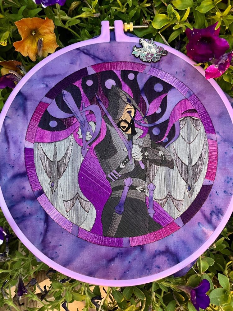 Photo of an embroidery hoop sitting on top of a flower pot. The fabric inside is purple with a partially completed embroidery of Lucanis from Dragon Age on it. The stitching is done in a style to somewhat mimic the appearance of stained glass.