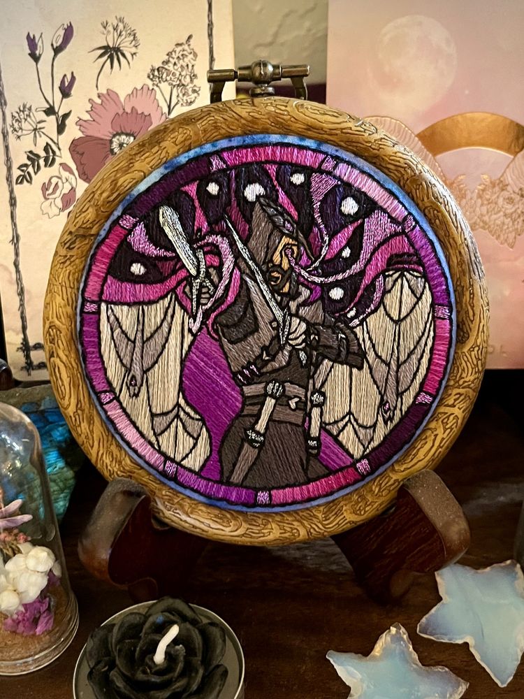 photograph of a small embroidery hoop sitting on a stand on a shelf, with small trinkets around it like tarot cards, a tea light candle, and moonstone/labradorite mineral hand specimens. 