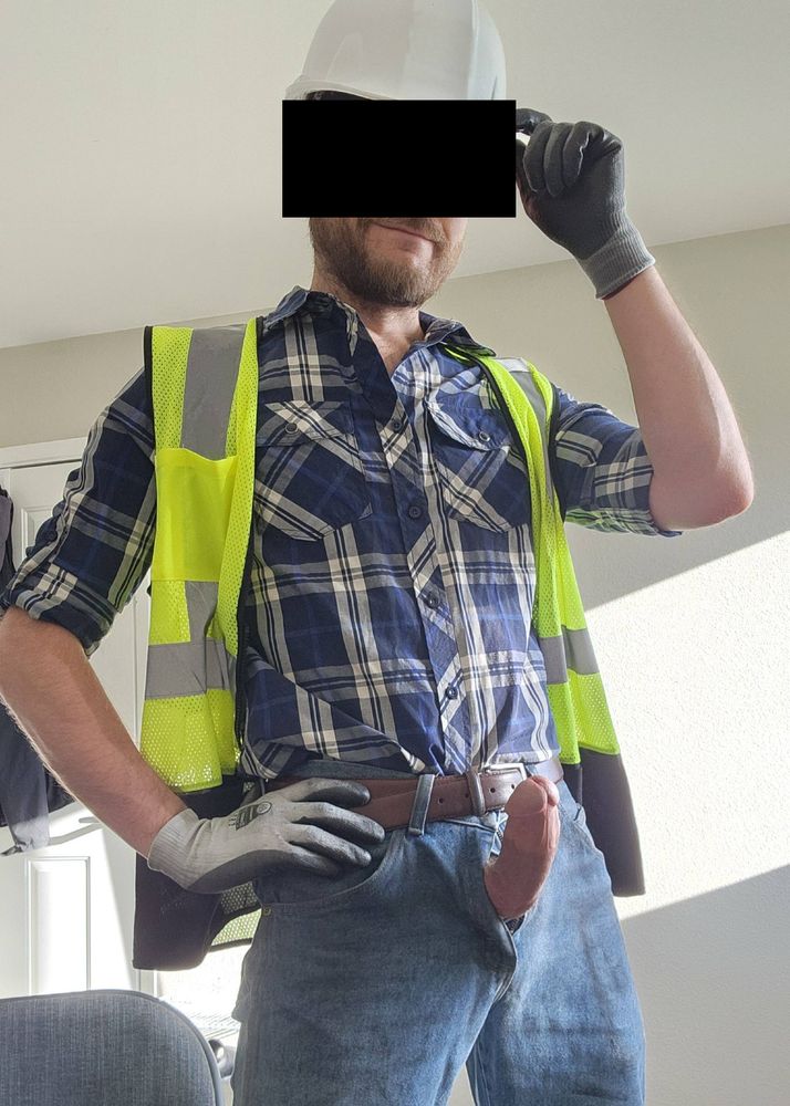 Myself wearing a blue plaid shirt under a green high-vis vest, beat up blue jeans, cut resistant gloves, and a white hard hat. The vest is open and my hard cock is protruding from my fly