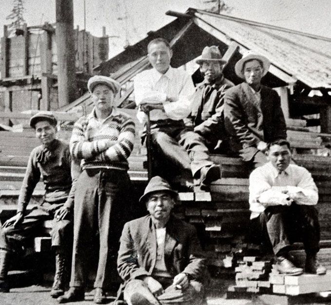 Seven Japanese men sitting and standing by a pile of lumber. 