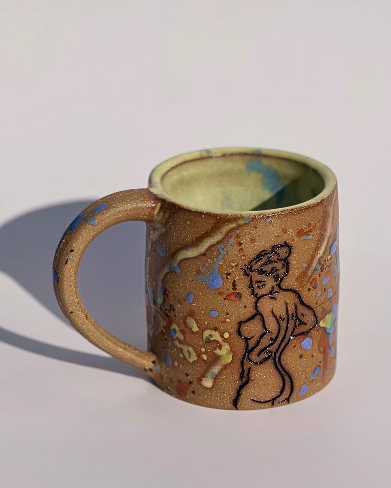 Handmade mug in brown stoneware decorated with a carved figure drawing and blue and yellow splattered glaze in front of a white background.