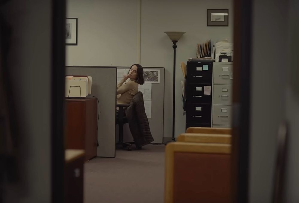 A frame from the movie “Sometimes I Think About Dying”. Daisy Ridley’s character is in a sterile office environment leaning back in here chair - looking off into the distance.