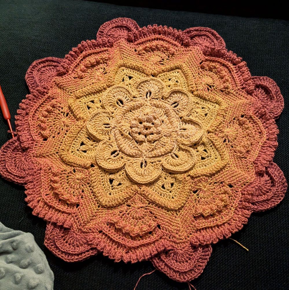 A work in progress crochet blanket, resembling a mandala. The yarn in the center of the circle is yellow and gradually changes to pink on the outside edges.