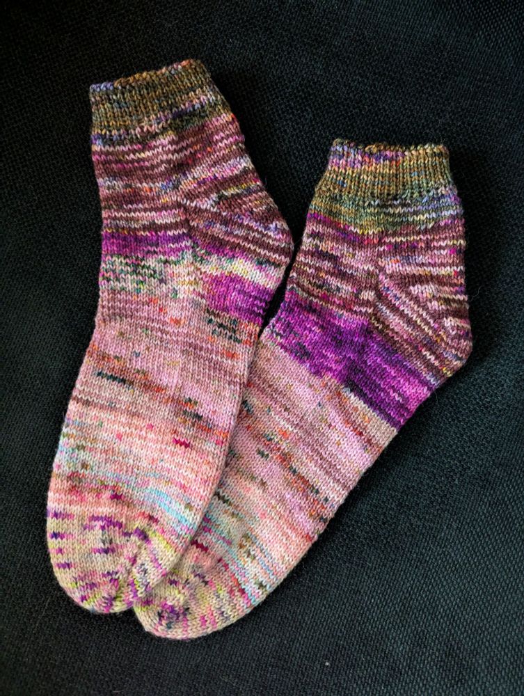 A pair of ankle length hand knitted socks, fading from variegated dark colors at the top to the toe. The toe is light pink and yellow, the heel is dark pink, and the cuff is green.