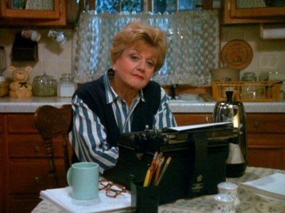 Jessica Fletcher from Murder She Wrote, played by Angela Lansbury, sits at her kitchen table in front of a typewriter. She looks at the camera, perhaps in a self satisfied way. There is no text.