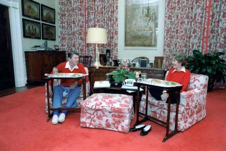 Nancy and Ronald Reagan on red patterned chairs with a red patterned ottoman and red patterned wallpaper