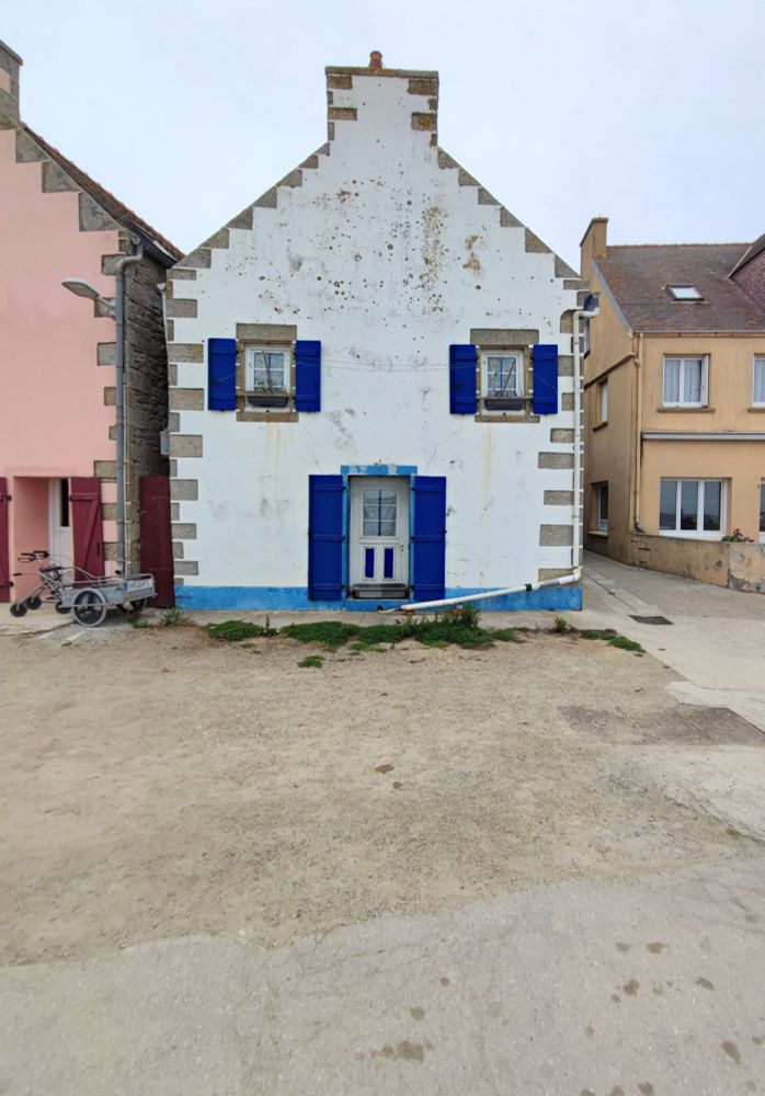 Ein weißes Haus mit zwei weißen Fenstern - eins rechts, eins links - und blauen Fensterläden im 1. OG und einer Tür mit blauen Läden mittig im Erdgeschoss.  