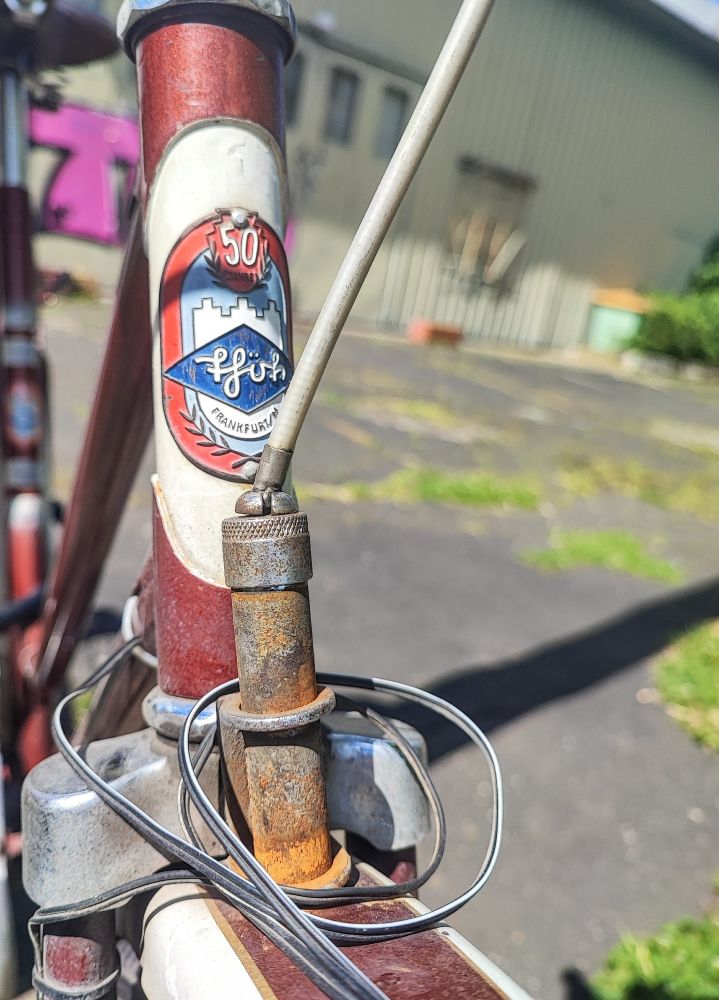 Das Bild zeigt den vorderen Teil eines  alten Fahrrads. Es ist das Steuerrohr mit einem Logo und die daran befestigten Komponenten, speziell der Bremszug, der von oben in die Aufnahme einer Stempelbremse führt zu sehen. Der Zug geht dabei nicht gerade verläuft dabei nicht gerade in die Aufnahme sondern sieht abgeknickt aus. 

Das Logo zeigt ein Schloss sowie die Aufschrift „50“ und „fföh Frankfurt/M.“. 