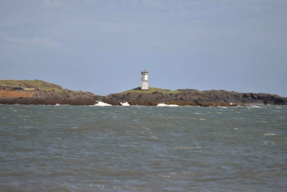 Meer, darüber hellblauer Himmel. Dazwischen ein Streifen braungrünes Land mit einem weißen Leuchtturm.