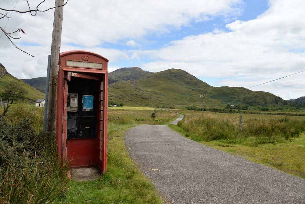 Eine rote britische, verwitterte Telefonzelle ohne Tür auf der linken Seite des Bilds. Rechts davon eine schmale Asphaltierte Straße, die durch grüne Wiesen führt. Im Hintergrund ein grüner Berg. 
