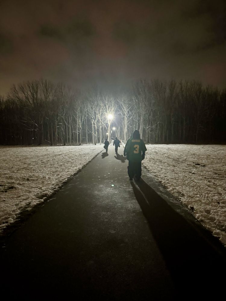 Notre Dame fans (one in a green #3 Montana jersey) walking on a well lit path cleared through a snowy field at night; snow covered trees are in the background.