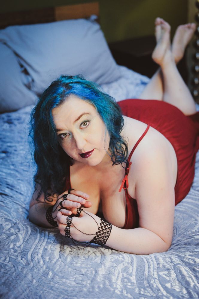 a curvy femme-presenting human woman lies facedown on a bed, propped up on her elbows, feet up, ankles crossed.  The bed is covered in a light blue velvet paisley duvet with pillows in light blue cases.  She's got long blue hair, red-painted lips, greenish eyes.  She's wearing a red slip that ties in ribbons on the shoulders, and black lace cuffs around her wrists that attach to black metal rings on each of her fingers, and each ring is connected to the cuff by a tiny chain.  She's holding her hands together and looking at the camera, and her cleavage is fantastic.  This photo was taken by Christy Shilling