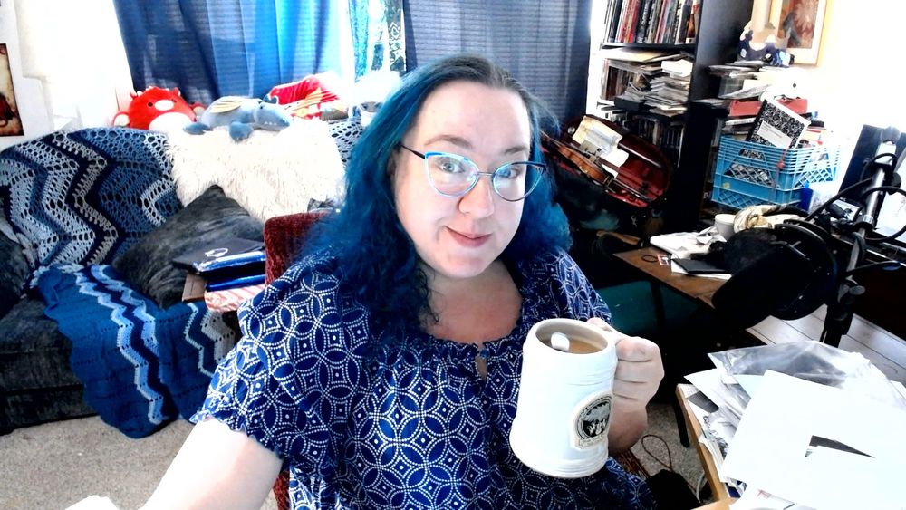 a curvy femme-presenting human lady holds up a tankard of tea with a spoon in it.  She's got long curly blue hair and blue iridescent cat-eye glasses, and she's wearing a blue shirt with a pattern of white interlocking circles and diamonds.  She's sitting in her living room, and in the room you can see her microphone, a violin, a bookshelf, and a couch with blue blankets and stuffed animals on it.  (Her favorite right now is the blue dragon with gold wings in the middle on top of the couch, but you can't tell that from the picture.)