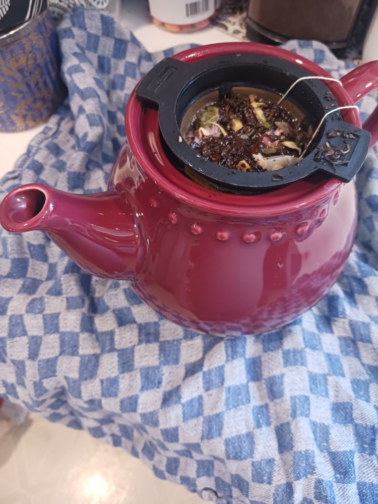 A big red teapot sits atop a grey and blue checkered dish towel. Tea leaves and rose buds are visible in a strainer basket.
