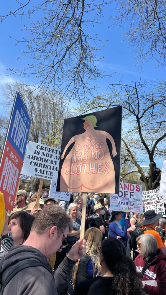 Protesters in the foreground with many signs, one showing the backside of Trump in comic format with the words, the emperor has no clothes. 