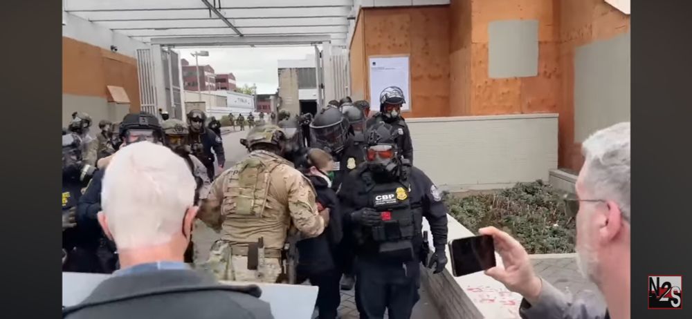 An ice agent in fatigues reaches through the other ice agents and grabs the young person, pulling them over the blue line and shoving them up the driveway towards the ice facility in Portland, Oregon on October 18, 2025