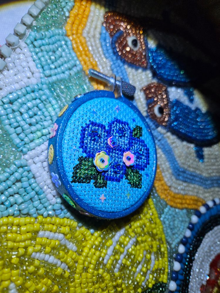 The blueberry themed cross stitch piece shown closer to show details, a 2.2" wooden embroidery hoop painted metallic blue and sprayed over with Mod Podge glitter sealer with alternating flower and leaf shaped spangles glued around the outside. The piece itself is stitched on blue 14 count iridescent aida cloth and uses metallic threads and glass seed beads with additional flower spangles stitched on the blueberries and iridescent sparkles glued about the front. Set against a beaded table runner in light colors.