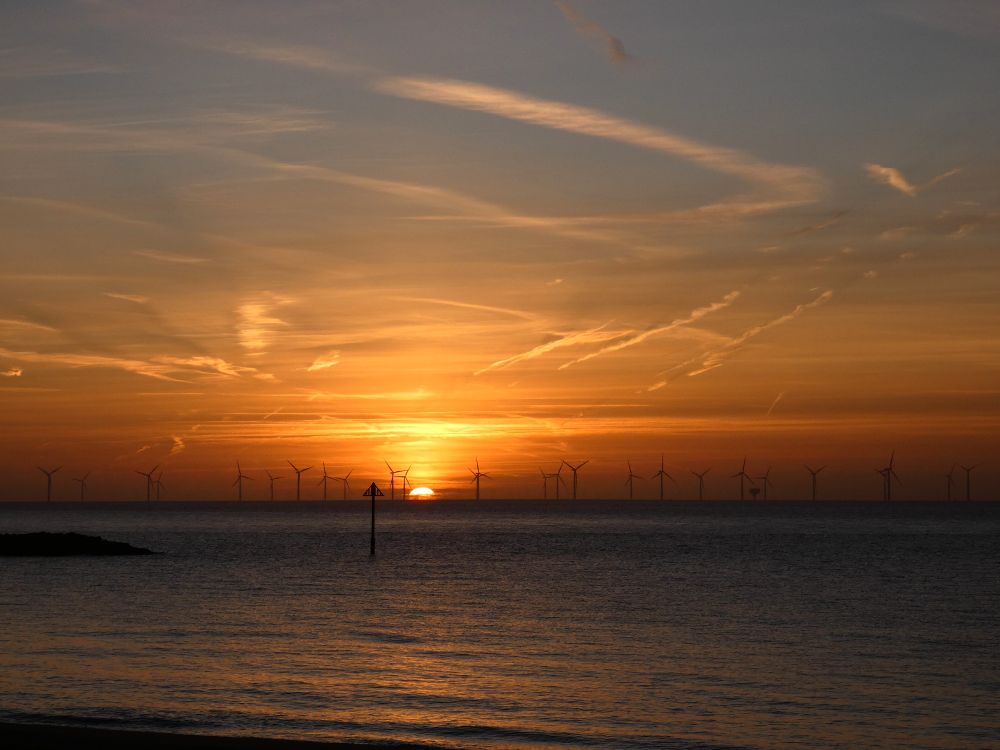 Silhouetted offshore wind turbines are visible against a vibrant orange and yellow sunrise over a dark sea.