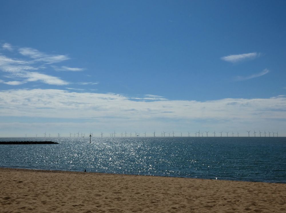 Sunny beach scene with a shimmering sea and a line of offshore wind turbines on the horizon.