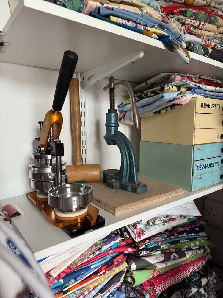 A badge and a button maker sit together on a shelf surrounded by folded fabric. 