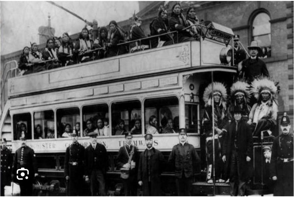An old black and white photograph of a Manchester tram with lots of native Americans in traditional clothes. There are police and conductors standing in front of the tram.