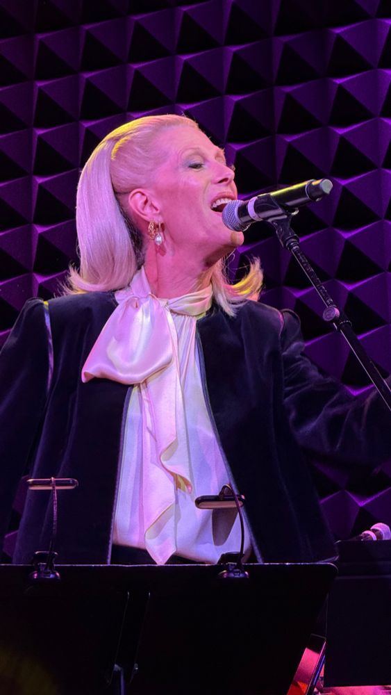Mx. Justin Vivian Bond performing at Joe's Pub in New York wearing a black and white suit lit brightly against a purple background