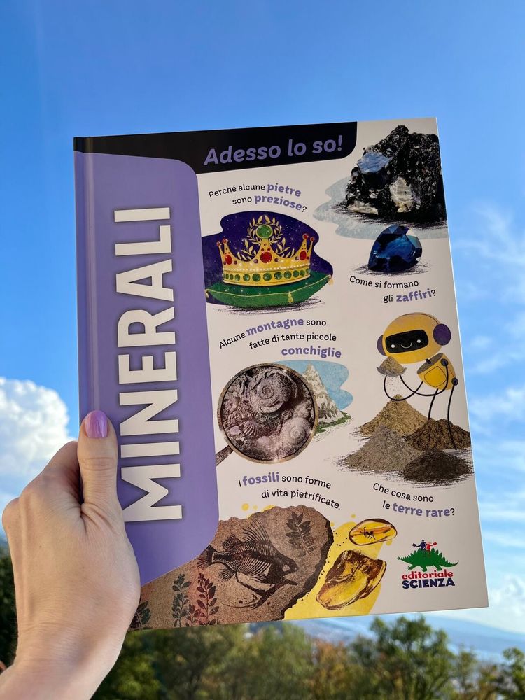 Una mano, in basso sinistra, sorregge un libro. La copertina del libro ha una fascia lilla a sinistra, con la scritta "Minerali", e sul resto della copertina ci sono delle illustrazioni di fossili, terre rare, una corona con gemme e uno zaffiro. Sullo sfondo della foto c'è un cielo azzurro e, alla base, degli alberi.